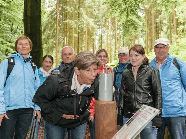 Gruppe von acht Personen in blauer und schwarzer Kleidung beim Waldausflug, eine Frau liest Infotafel.