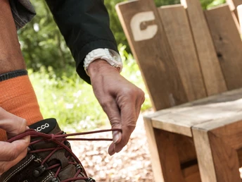 Person bindet braune Wanderstiefel vor einer hölzernen Sitzbank in Waldumgebung.