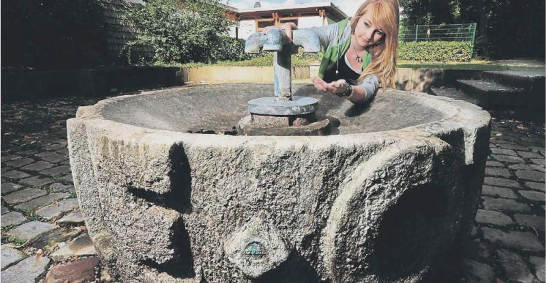 Steinbrunnen mit historischer Ornamentik, Frau betätigt den Wasserhahn, um frisches Quellwasser aufzufangen.