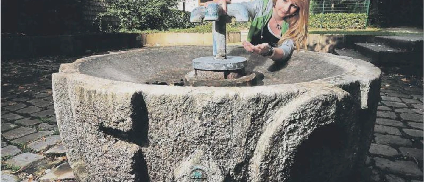 Steinbrunnen mit historischer Ornamentik, Frau betätigt den Wasserhahn, um frisches Quellwasser aufzufangen.