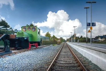 Eine historische grüne Geha-Lok steht auf Schienen am Hövelhofer Bahnhof, eingerahmt von Bäumen.