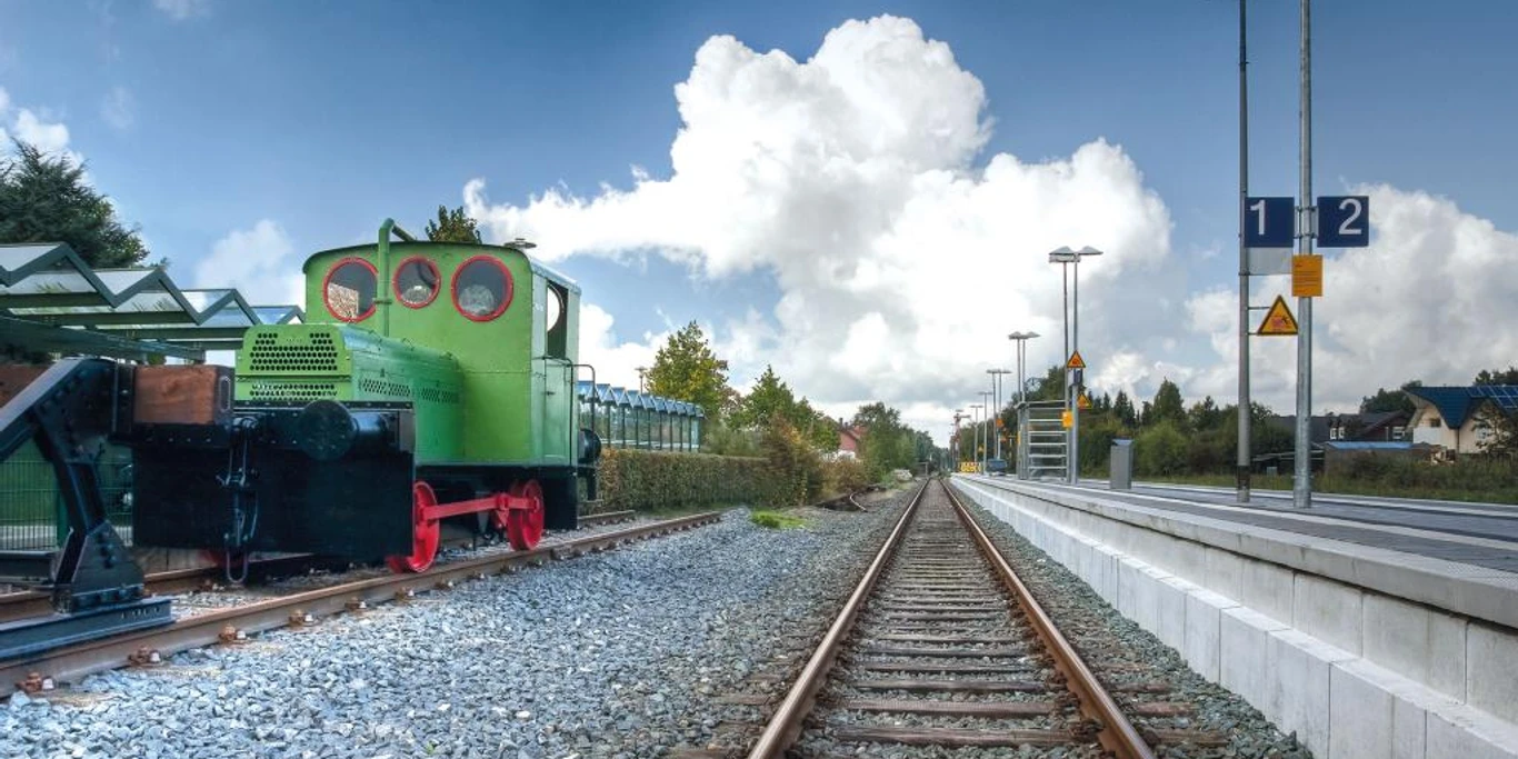 Eine historische grüne Geha-Lok steht auf Schienen am Hövelhofer Bahnhof, eingerahmt von Bäumen.