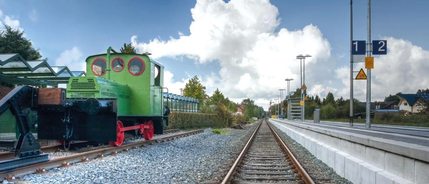 Eine historische grüne Geha-Lok steht auf Schienen am Hövelhofer Bahnhof, eingerahmt von Bäumen.