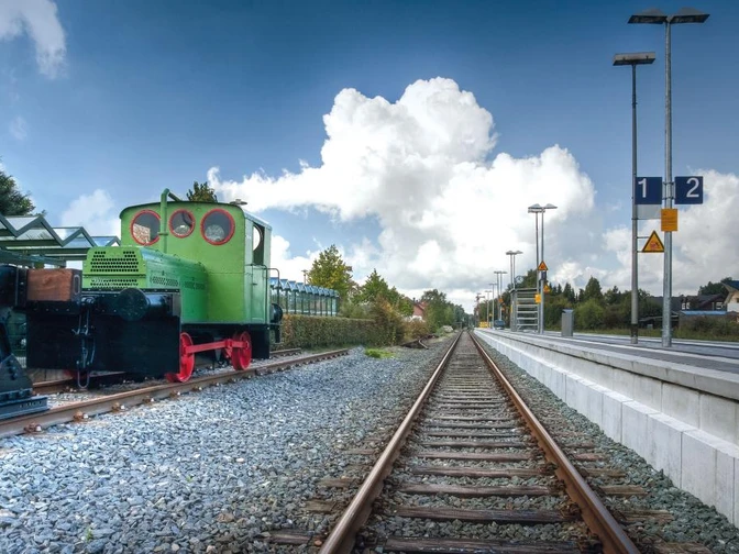 Eine historische grüne Geha-Lok steht auf Schienen am Hövelhofer Bahnhof, eingerahmt von Bäumen.