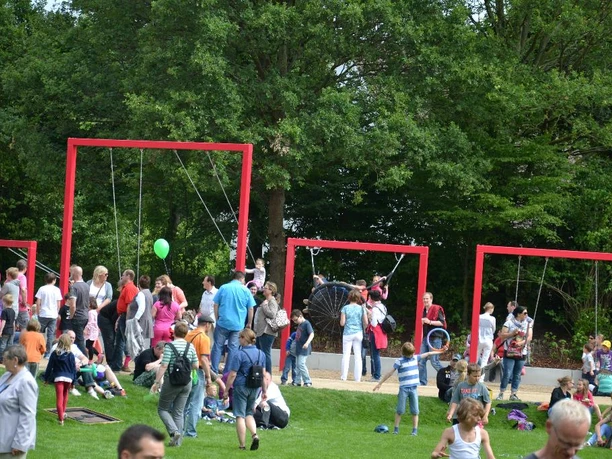 Menschen im Park genießen ein sonniges Fest mit roten Rahmen-Schaukeln, umgeben von Bäumen.