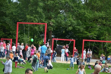 Schaukelparade im Flora-Westfalica-Park, Rheda-Wiedenbrück Menschen im Park genießen ein sonniges Fest mit roten Rahmen-Schaukeln, umgeben von Bäumen.
