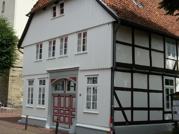 Storchenhaus in NRÜ Außenansicht Fachwerkhaus mit rotem Ziegeldach und weißen Fenstern, teils verschattet unter blauem Himmel.
