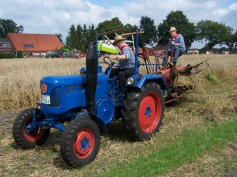 Aperberg Zwei Männer mit Strohhüten mähen Korn auf dem Feld mit einem blauen Oldtimer-Traktor.corn mowing