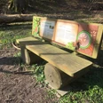 Holzbank mit Rückenlehne auf Wiese, umgeben von Bäumen. Informationstafel zum Naturlehrpfad.