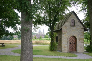 Hovekapelle Eine kleine steinerne Kapelle steht malerisch zwischen hohen Bäumen, mit zwei Kirchtürmen im Hintergrund.