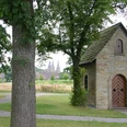 Hovekapelle Eine kleine steinerne Kapelle steht malerisch zwischen hohen Bäumen, mit zwei Kirchtürmen im Hintergrund.