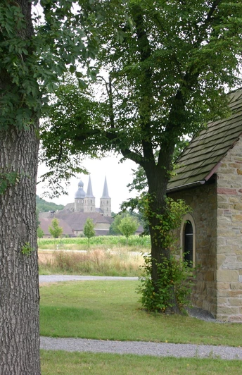 Hovekapelle Eine kleine steinerne Kapelle steht malerisch zwischen hohen Bäumen, mit zwei Kirchtürmen im Hintergrund.