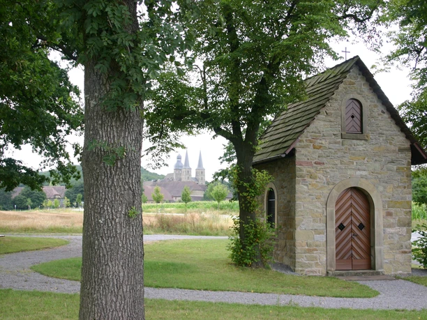 Hovekapelle Eine kleine steinerne Kapelle steht malerisch zwischen hohen Bäumen, mit zwei Kirchtürmen im Hintergrund.