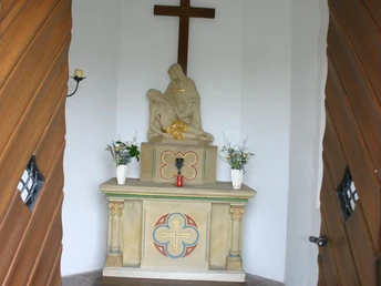 Hovekapelle (Innenansicht) Blick durch schwere Holztüren auf einen schlichten Altar mit Marmorskulptur und Kreuz im Hintergrund.