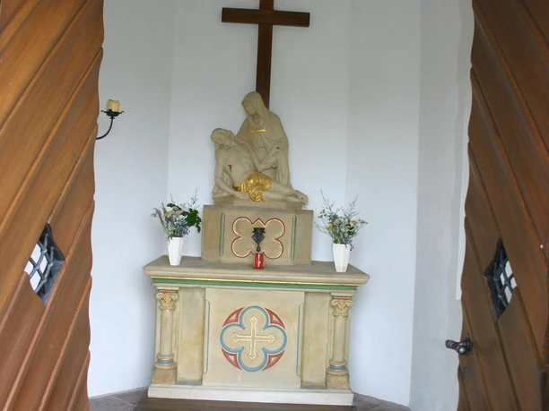 Hovekapelle (Innenansicht) Blick durch schwere Holztüren auf einen schlichten Altar mit Marmorskulptur und Kreuz im Hintergrund.