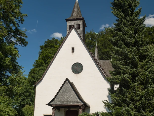 Die Kapelle der Malteser Komturei <p>Historische Kirche mit spitzem Turm, umgeben von üppigen Bäumen und blühenden Sträuchern.</p>