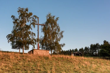 Das Osterkreuz Ein großes Holzkreuz neben zwei Bäumen auf einem grasbewachsenen Hügel vor klarem, blauem Himmel.