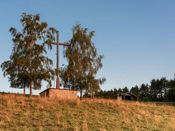 Das Osterkreuz Ein großes Holzkreuz neben zwei Bäumen auf einem grasbewachsenen Hügel vor klarem, blauem Himmel.