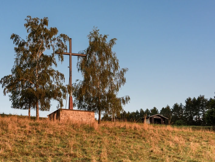 Das Osterkreuz Ein großes Holzkreuz neben zwei Bäumen auf einem grasbewachsenen Hügel vor klarem, blauem Himmel.