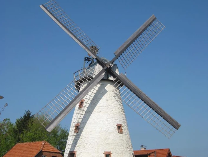 Historische Windmühle mit weißem Turm und Holzelementen, umgeben von Häusern und Bäumen unter blauem Himmel.