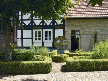 Historisches Fachwerkgebäude mit gepflegtem Garten und kunstvollem Mosaik im Vordergrund.