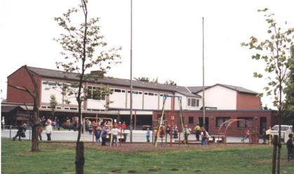 Kinder spielen auf einem Spielplatz vor einer Schule mit rot-weißen Gebäuden im Hintergrund.