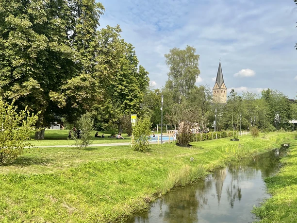 Wiehlpark Ein idyllischer Park mit Bach, grüner Wiese und Kirchturm im Hintergrund unter wolkigem Himmel.
