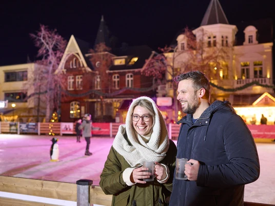 Bei einem Heißgetränk die Eisbahn genießen Paar genießt heißen Glühwein vor festlich beleuchteter Eisbahn, umgeben von historischen Gebäuden.