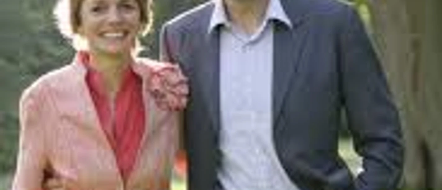 Ein lächelndes Paar spaziert in formeller Kleidung durch einen grünen Park bei Sonnenlicht.