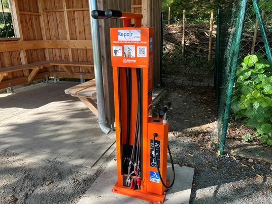 Fahrrad-Reparaturstation Leuchtend orangefarbene Fahrrad-Reparaturstation mit Werkzeugen und Luftpumpe, befestigt an einem überdachten Rastplatz nahe einem Waldweg.