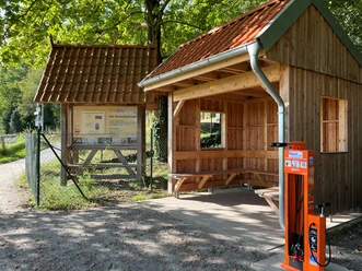Fahrrad-Reparaturstation neben hölzerner Unterstandshütte und Infotafel, umgeben von Bäumen.