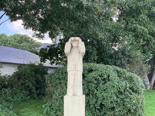 Mann mit Fernglas Steinfigur eines Mannes mit Fernglas auf hohem Sockel in grüner Parklandschaft, umrahmt von Büschen.