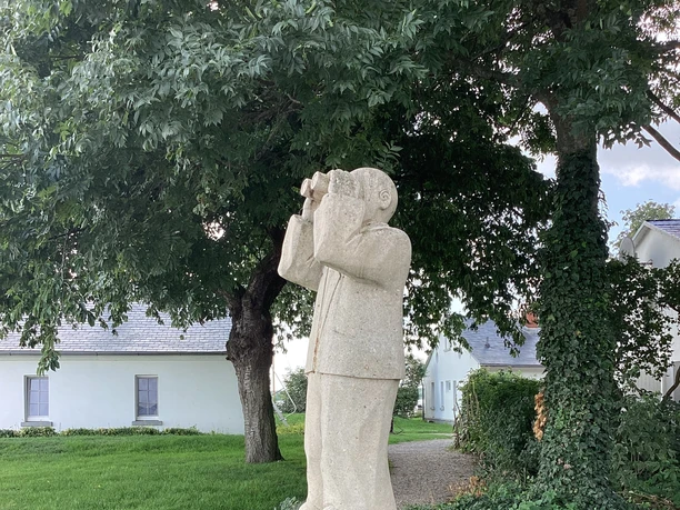 Steinfigur eines Mannes mit Fernglas im Grünen, die in die Ferne blickt, umgeben von Bäumen.