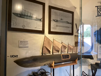 Ein historisches Modell eines Segelschiffs in einem Museum, mit Zeichnungen darüber an der Wand.