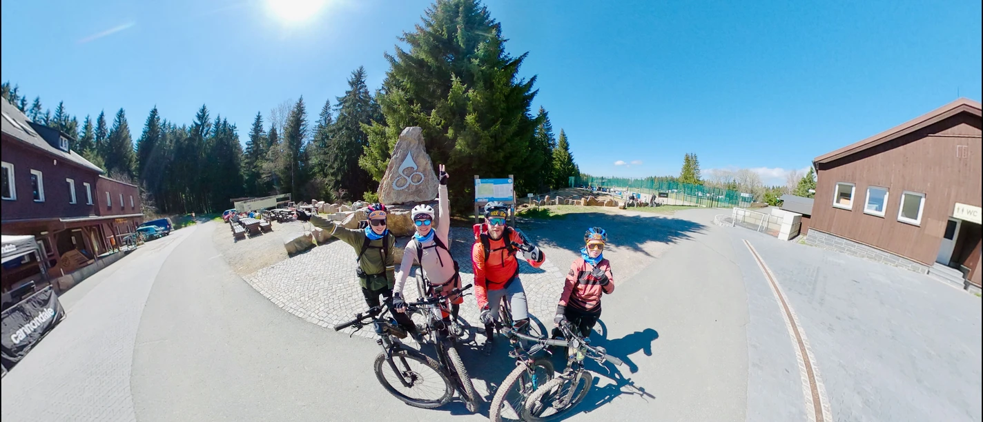 1_Stoneman Miriquidi (c) TMGS_Fravely.jpeg Vier Radfahrer posieren mit Mountainbikes auf einem sonnigen Platz vor einem Holzhäuserensemble.