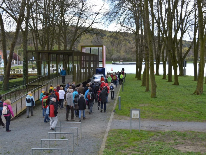 Eine Gruppe von Wanderern gelangt über einen schmalen Gehweg zu einer überdachten Info-Box am See.