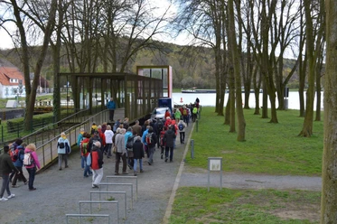 Info-Box am Naxxen Dreieck in Hörstel Eine Gruppe von Wanderern gelangt über einen schmalen Gehweg zu einer überdachten Info-Box am See.