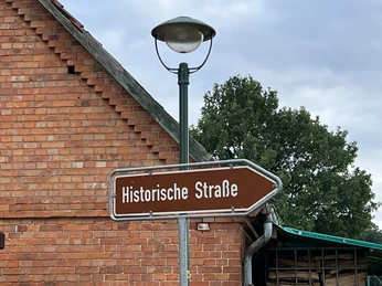 Altes Straßenschild mit der Aufschrift "Historische Straße" vor einer Ziegelmauer und Bäumen.
