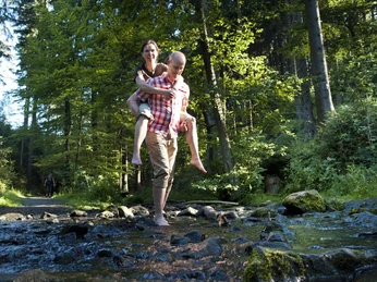 Ein Mann trägt eine Frau auf dem Rücken, während beide barfuß einen flachen Bach durchqueren.