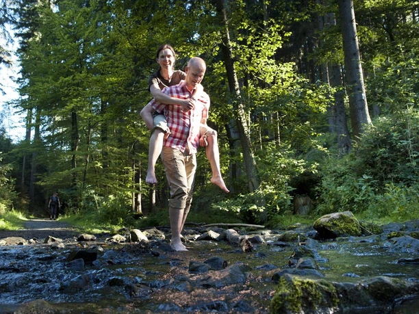 Spaß im Silberbachtal Ein Mann trägt eine Frau auf dem Rücken, während beide barfuß einen flachen Bach durchqueren.