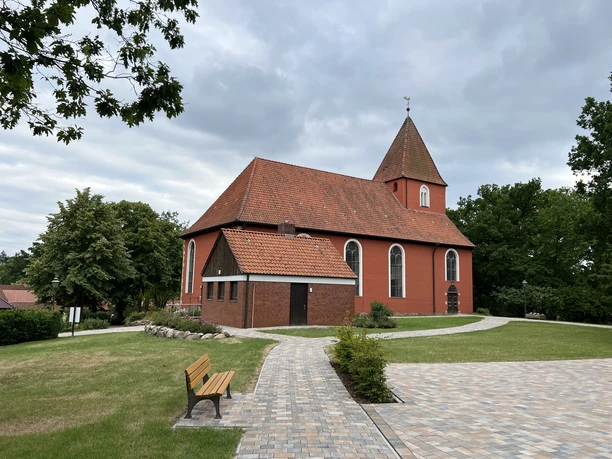 St. Petri-Kirche Kirchlinteln Braune Backsteinkirche mit rotem Dach, umgeben von Bäumen, vor bewölktem Himmel in ruhiger Umgebung.