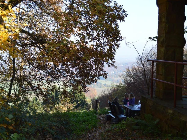 Herbstliche Szene mit weitem Blick ins Tal, Parkbänke und Besucher unter farbenfrohen Baumkronen.