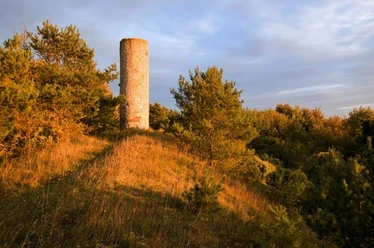 Ein runder Steinturm steht umgeben von grünem Laubwerk auf einem Hügel bei warmem Abendlicht.