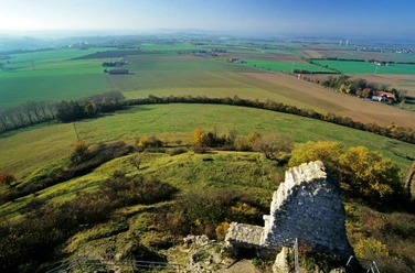 Ruinenmauer auf Grünhügel vor weiten, sanft getönten Feldern und entfernten Horizontlinien.