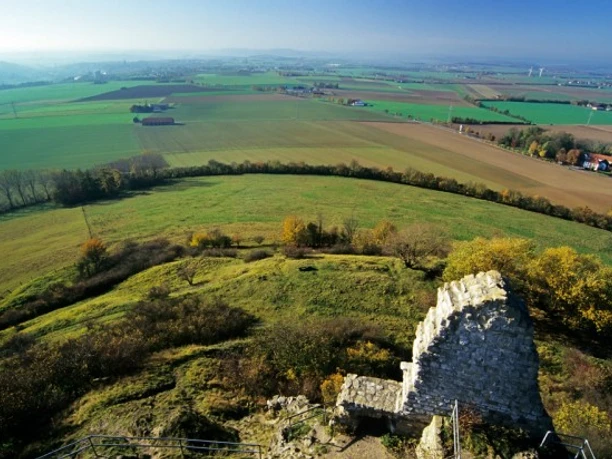 Ruinenmauer auf Grünhügel vor weiten, sanft getönten Feldern und entfernten Horizontlinien.