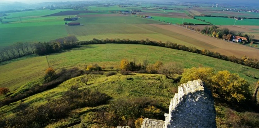Ruinenmauer auf Grünhügel vor weiten, sanft getönten Feldern und entfernten Horizontlinien.