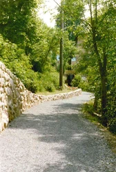 Wanderweg hinter der Mühle Schotterweg führt sanft geschwungen durch grüne Bäume, Mühle sichtbar im Hintergrund, sonniger Tag.