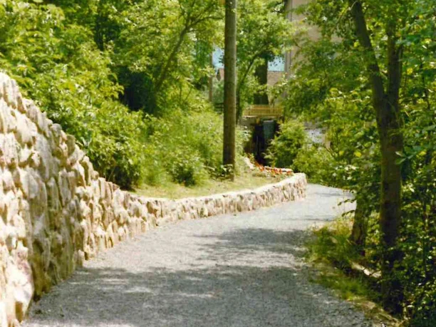 Wanderweg hinter der Mühle Schotterweg führt sanft geschwungen durch grüne Bäume, Mühle sichtbar im Hintergrund, sonniger Tag.