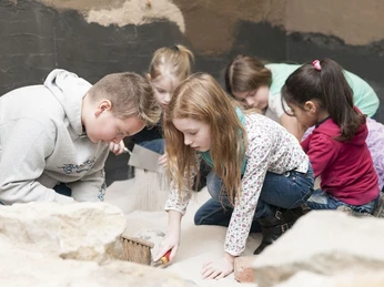 Kinder erforschen gemeinsam einen Sandkasten, konzentriert auf einen archäologischen Ausgrabungsnachbau.