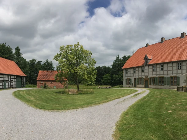 Historische Fachwerkgebäude unter bewölktem Himmel, eingebettet in grüne Wiesen und Bäume.